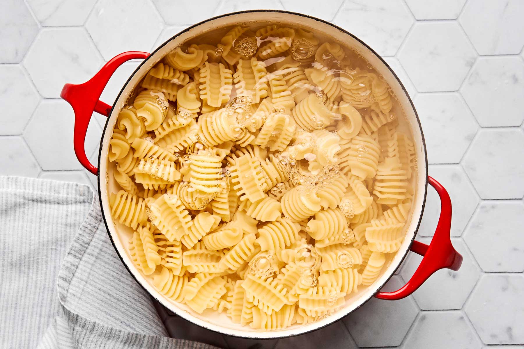 A red pot filled with boiling water and radiatori pasta sits on a white hexagon-tile countertop next to a striped kitchen towel.