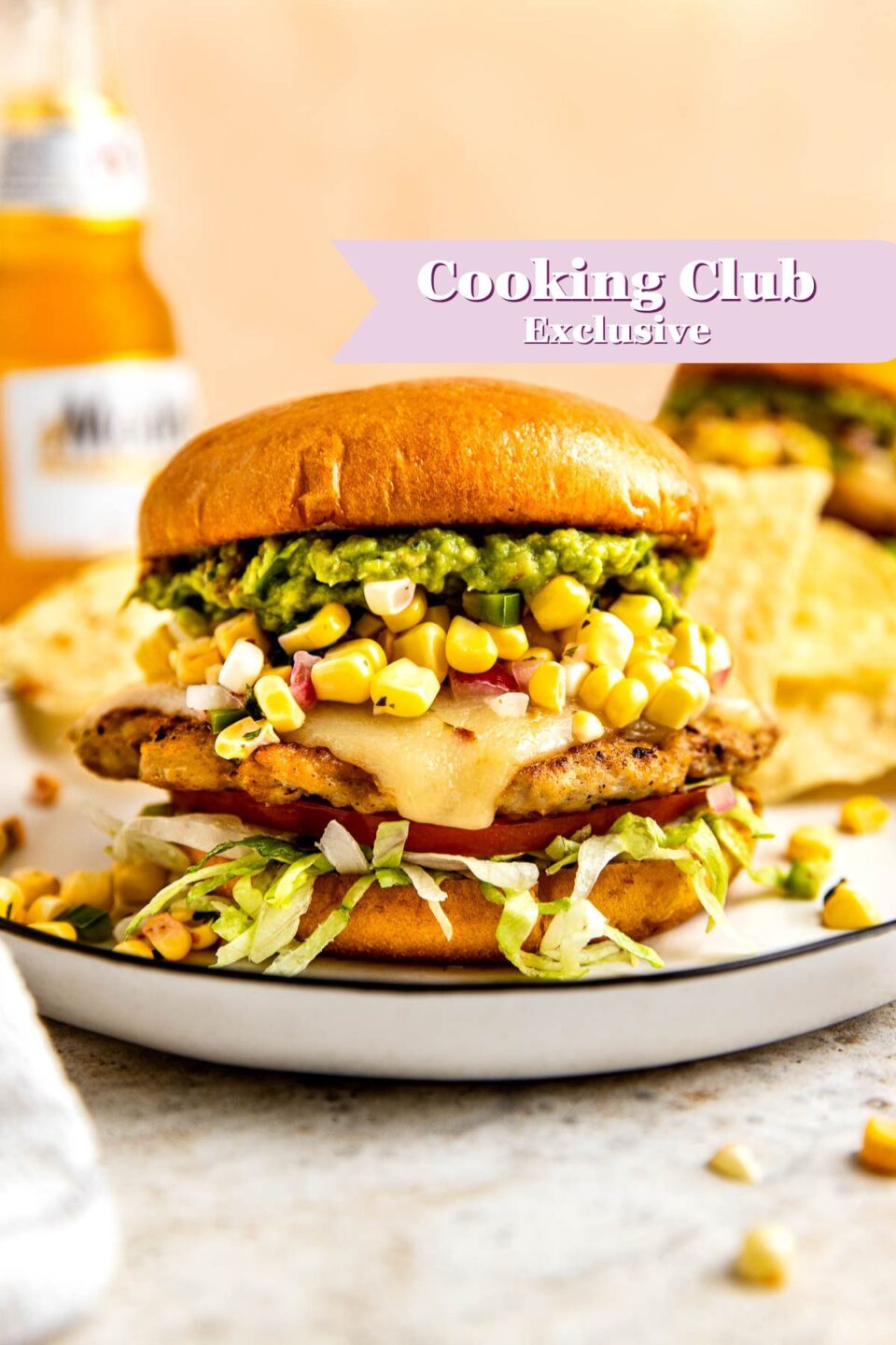 A grilled chicken sandwich with melted cheese, lettuce, corn salsa, and guacamole on a toasted bun sits on a plate. A beer bottle and tortilla chips are in the background. Text reads Cooking Club Exclusive.
