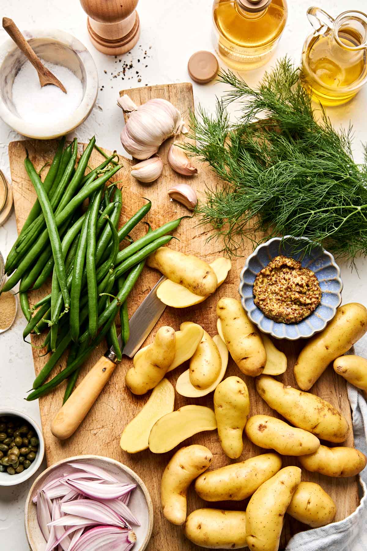 Fingerling potatoes, green beans, garlic, fresh dill, olive oil, mustard, capers, shallots, salt, and pepper are arranged on a wooden board and white surface, ready for cooking or salad preparation.