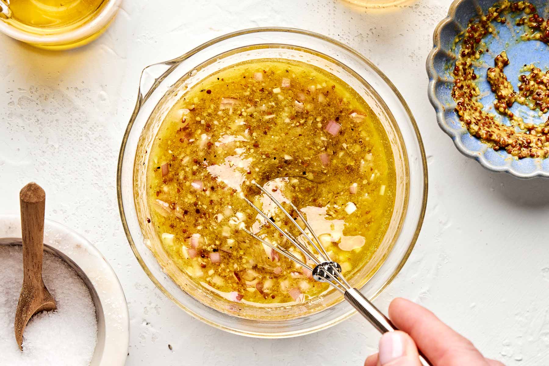 A hand uses a small whisk to mix a bowl of vinaigrette with chopped shallots, herbs, and black pepper; salt and mustard are in bowls nearby on a white surface.