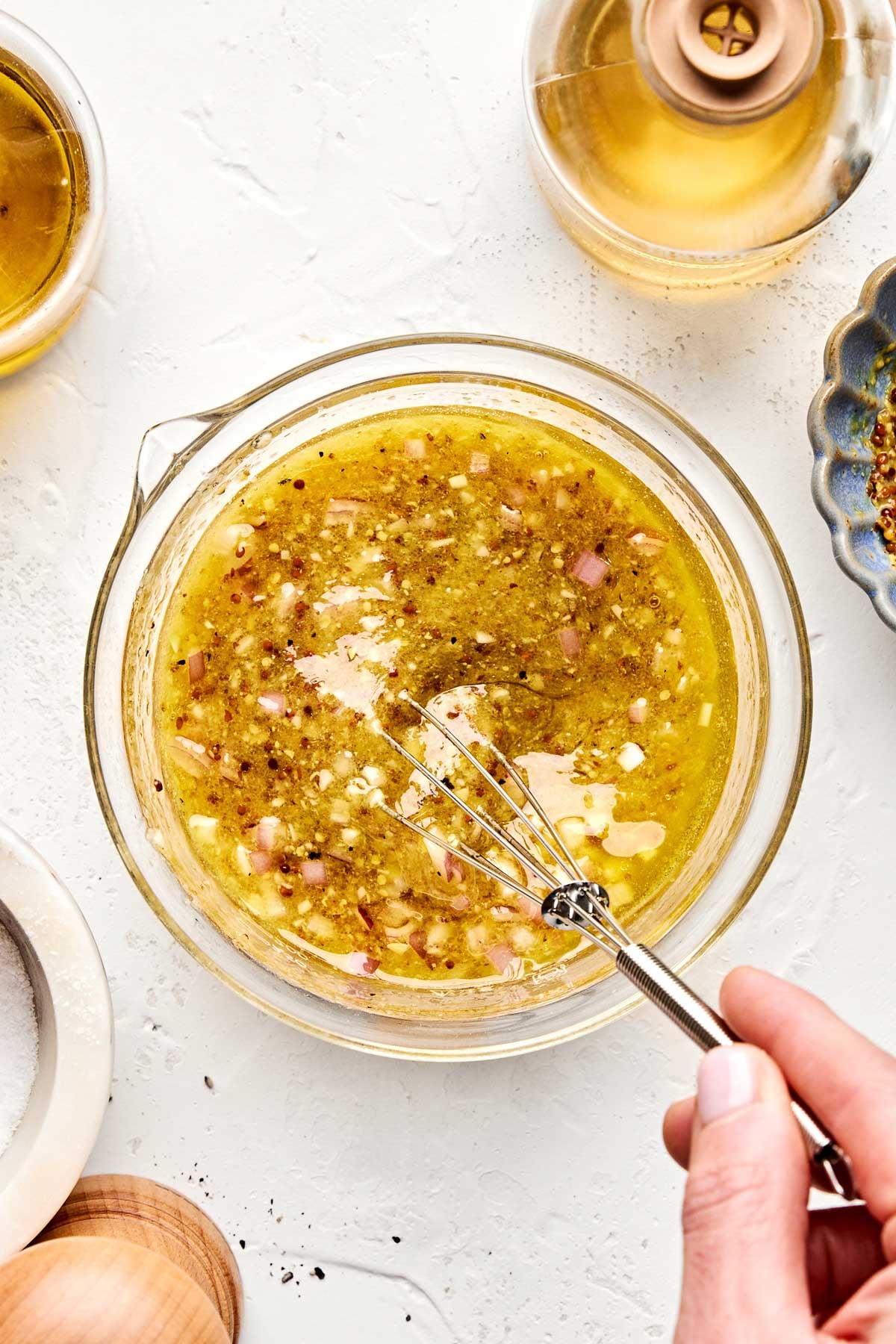 A hand holds a whisk, mixing an olive oil-based vinaigrette with minced shallots and seasonings in a glass bowl on a white countertop, surrounded by bottles and bowls.