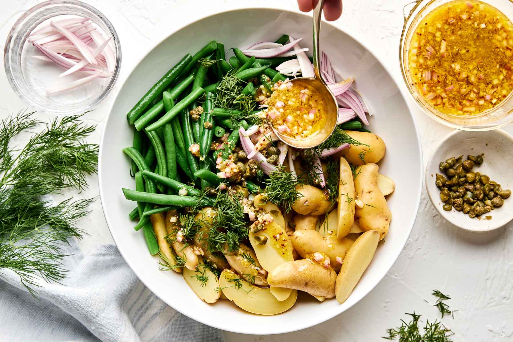 A bowl with sliced potatoes, green beans, fresh dill, and red onion, as vinaigrette is poured over. Capers and extra vinaigrette are nearby, with a glass of water and a napkin on a white table.