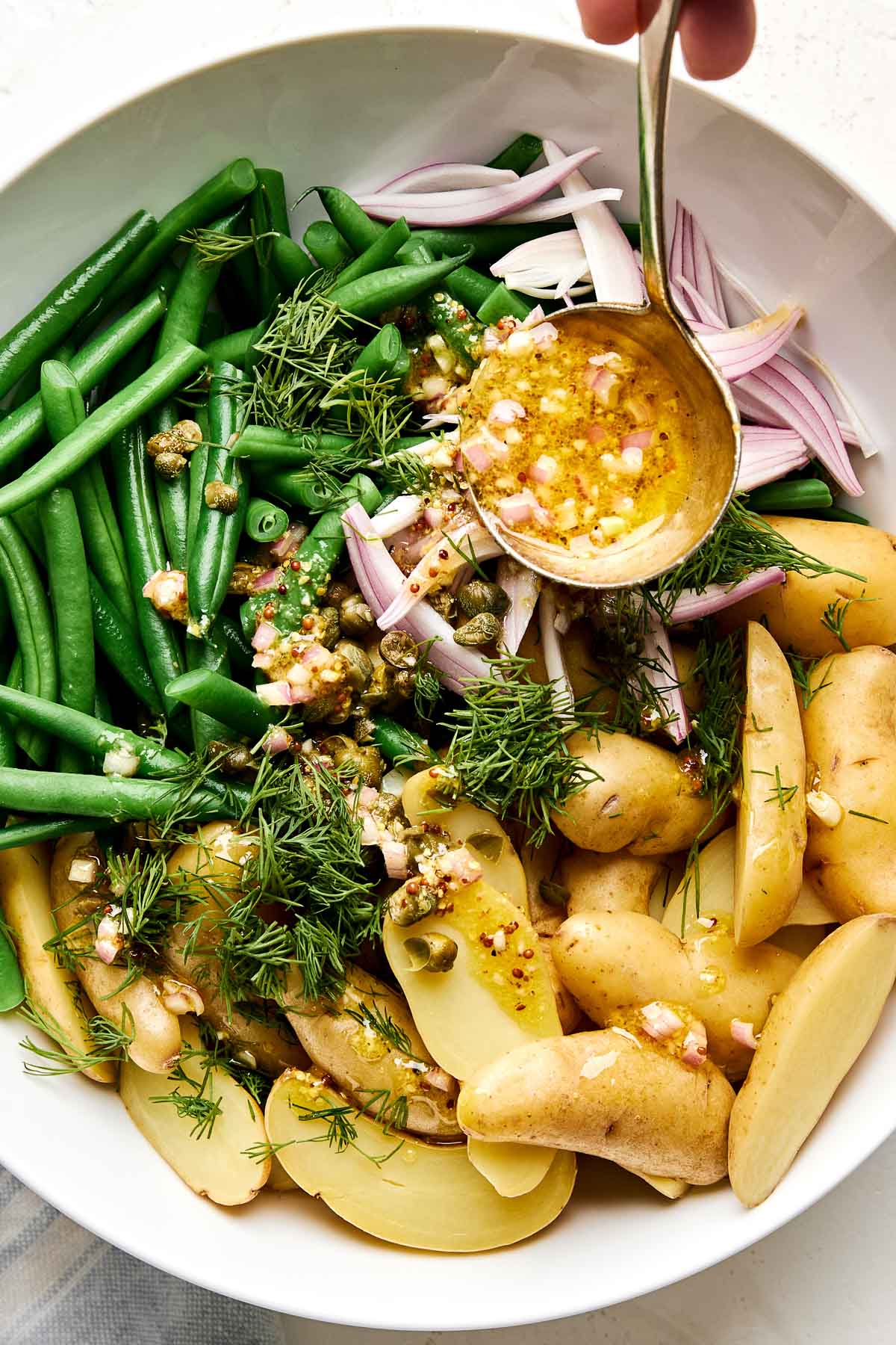 A bowl filled with green beans, sliced potatoes, capers, fresh dill, and sliced shallots, with a hand pouring a mustard vinaigrette over the vegetables.