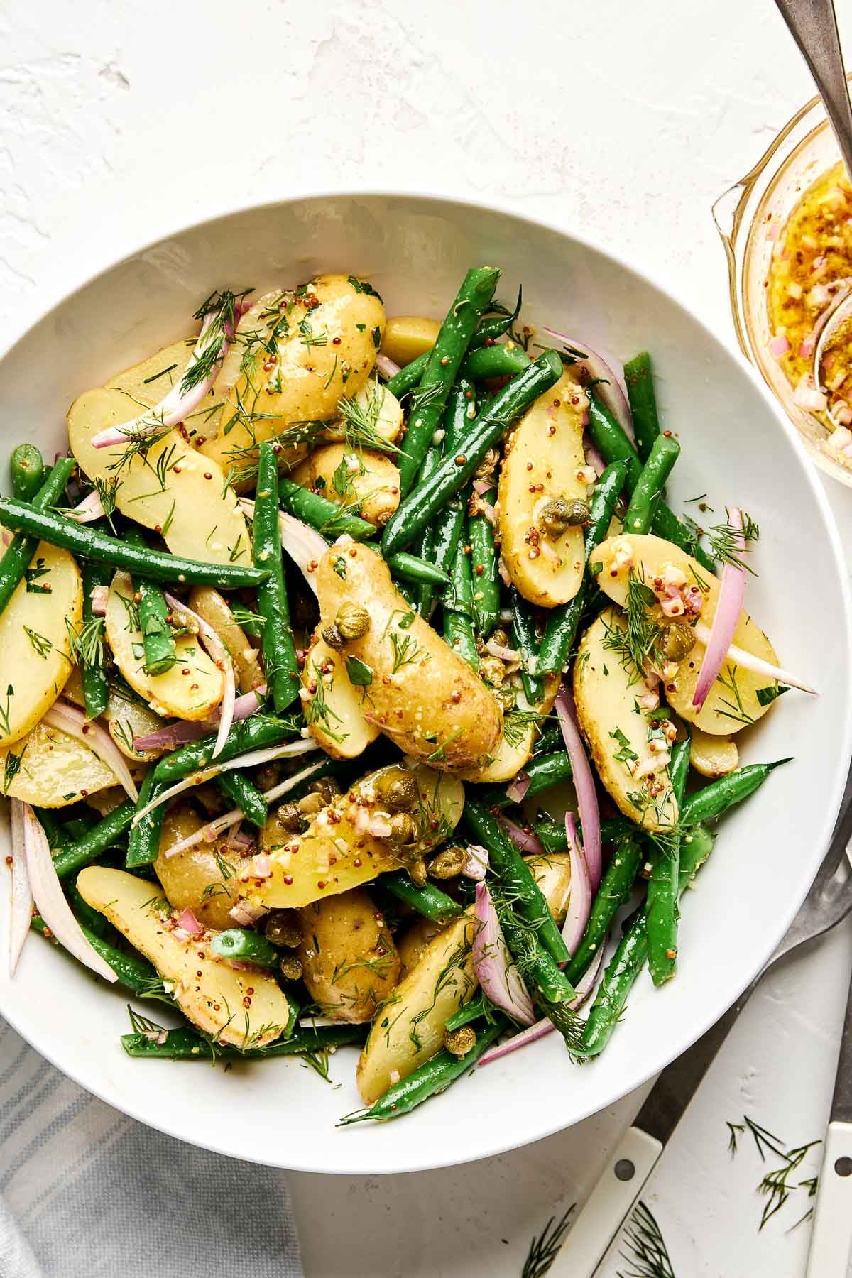 A bowl of potato salad with fingerling potatoes, green beans, sliced shallots, capers, and fresh dill, all tossed in a light vinaigrette. A fork and a small bowl of mustard dressing are nearby.