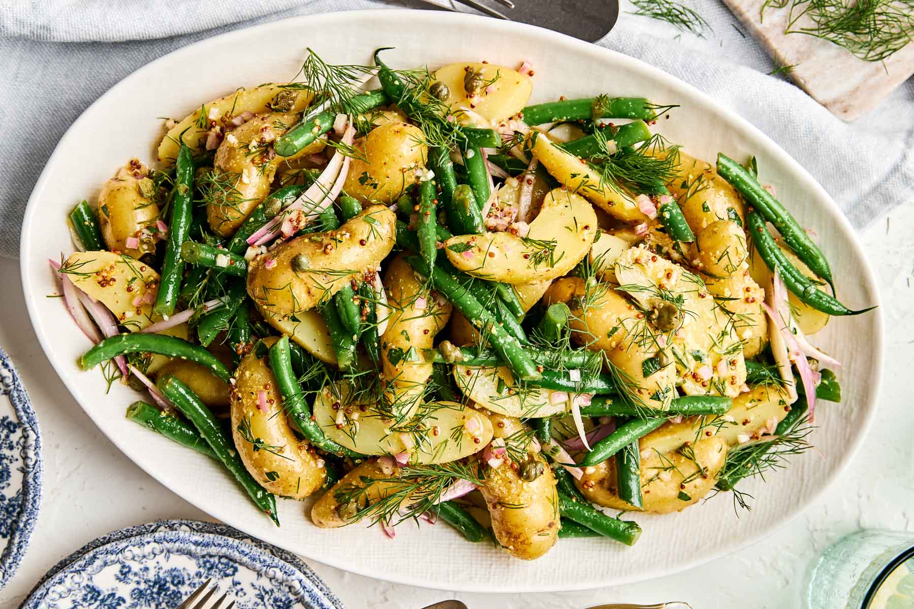 A serving platter filled with potato salad featuring fingerling potatoes, green beans, red onions, chopped dill, and a grainy mustard dressing, garnished with fresh herbs.