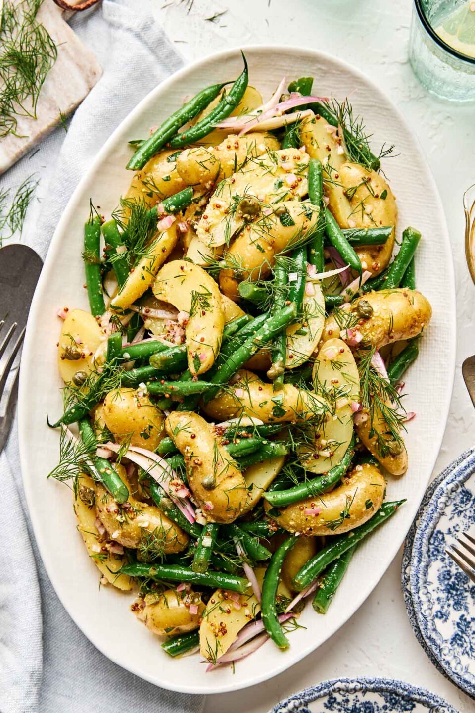 An oval platter filled with potato salad made of fingerling potatoes, green beans, shallots, and fresh dill, tossed in a mustard vinaigrette. The dish is set on a light-colored table with plates and cutlery nearby.