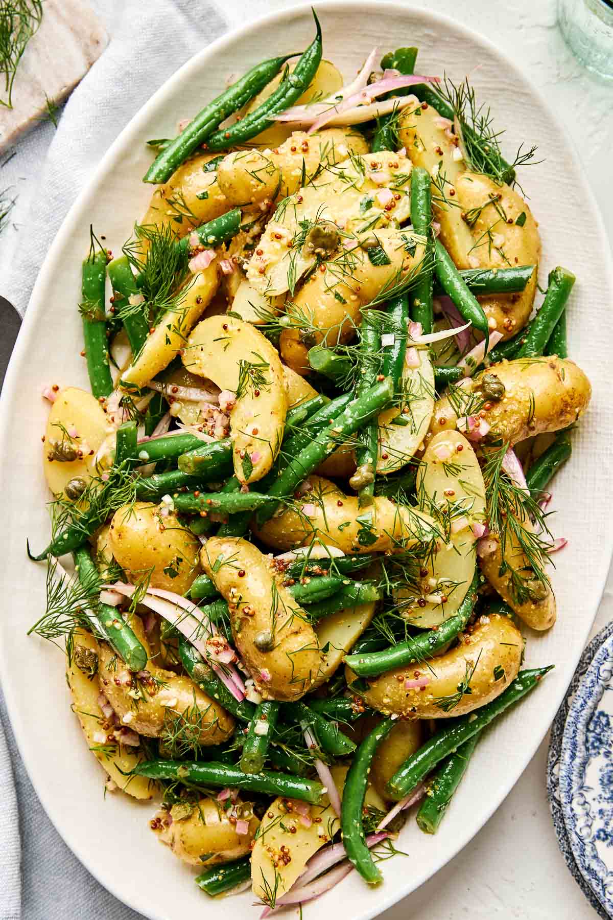 An oval platter filled with a potato salad made of sliced potatoes, green beans, shallot, capers, and fresh dill, dressed with a grainy mustard vinaigrette.