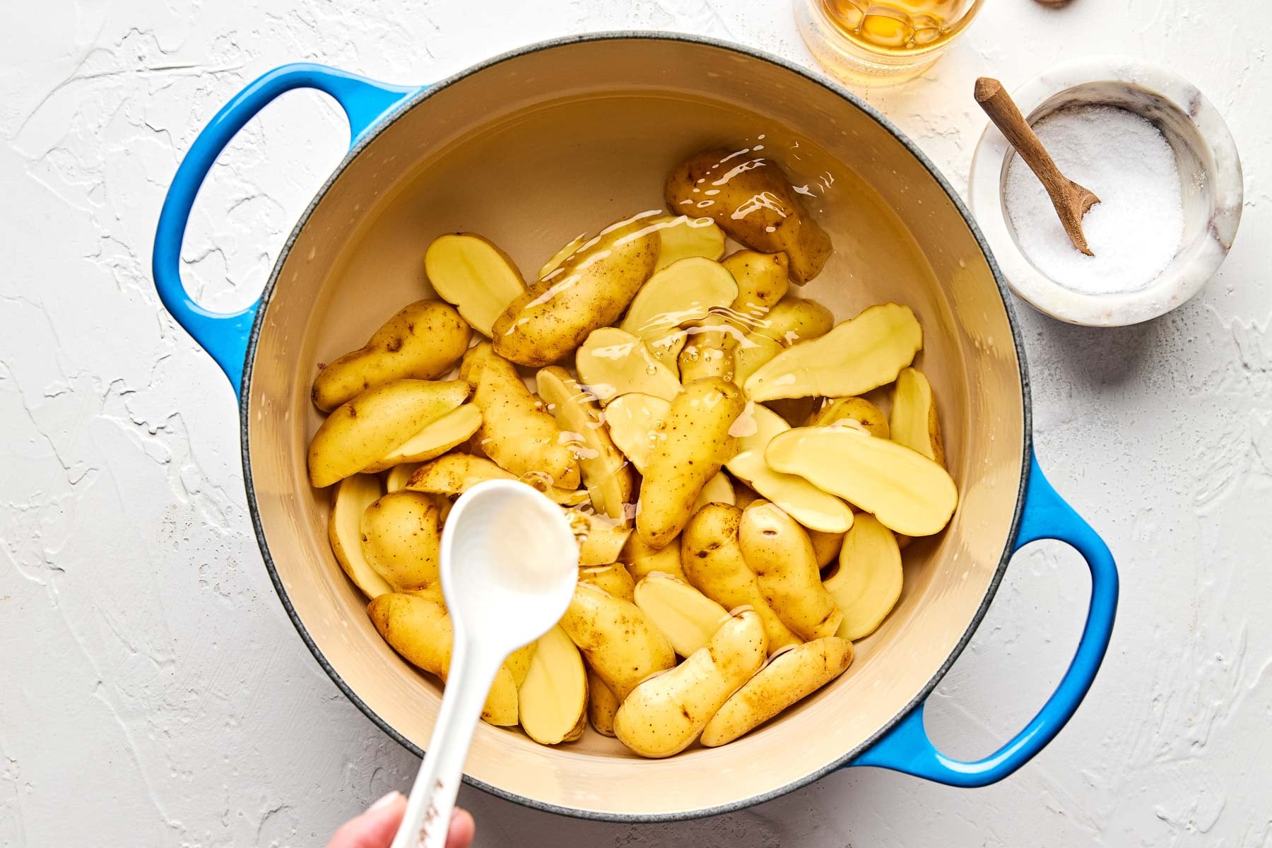 A blue pot filled with fingerling potatoes, some whole and some sliced, sits on a white surface with water being poured in. Nearby are a small bowl of salt and a container of oil. A hand holds a white spoon over the pot.