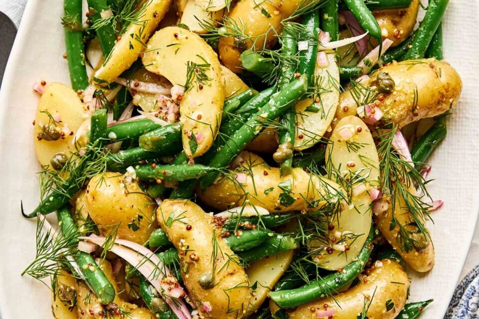 A close-up of a potato salad with fingerling potatoes, green beans, sliced red onions, capers, chopped herbs, and whole grain mustard dressing on a white plate.