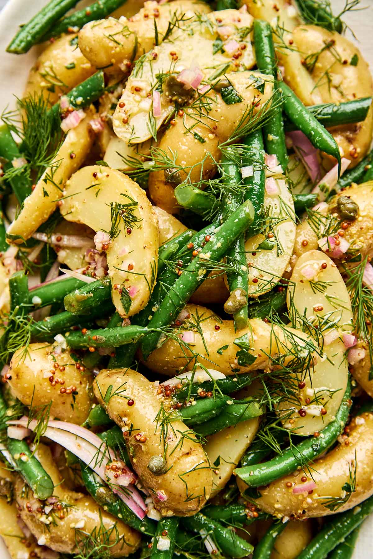 Close-up of a potato salad with green beans, red onion slices, fresh dill, capers, and whole grain mustard dressing. The ingredients are mixed together, showcasing vibrant green, yellow, and pink colors.