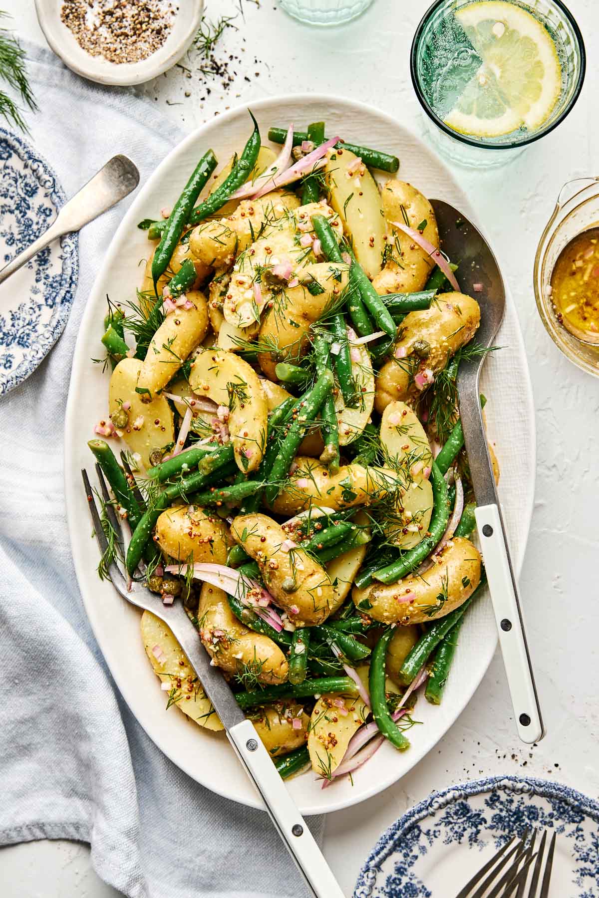A white oval platter filled with potato salad featuring fingerling potatoes, green beans, red onions, and fresh dill, with salad servers. A lemon drink, dressing jar, and seasoning are nearby on a white table.