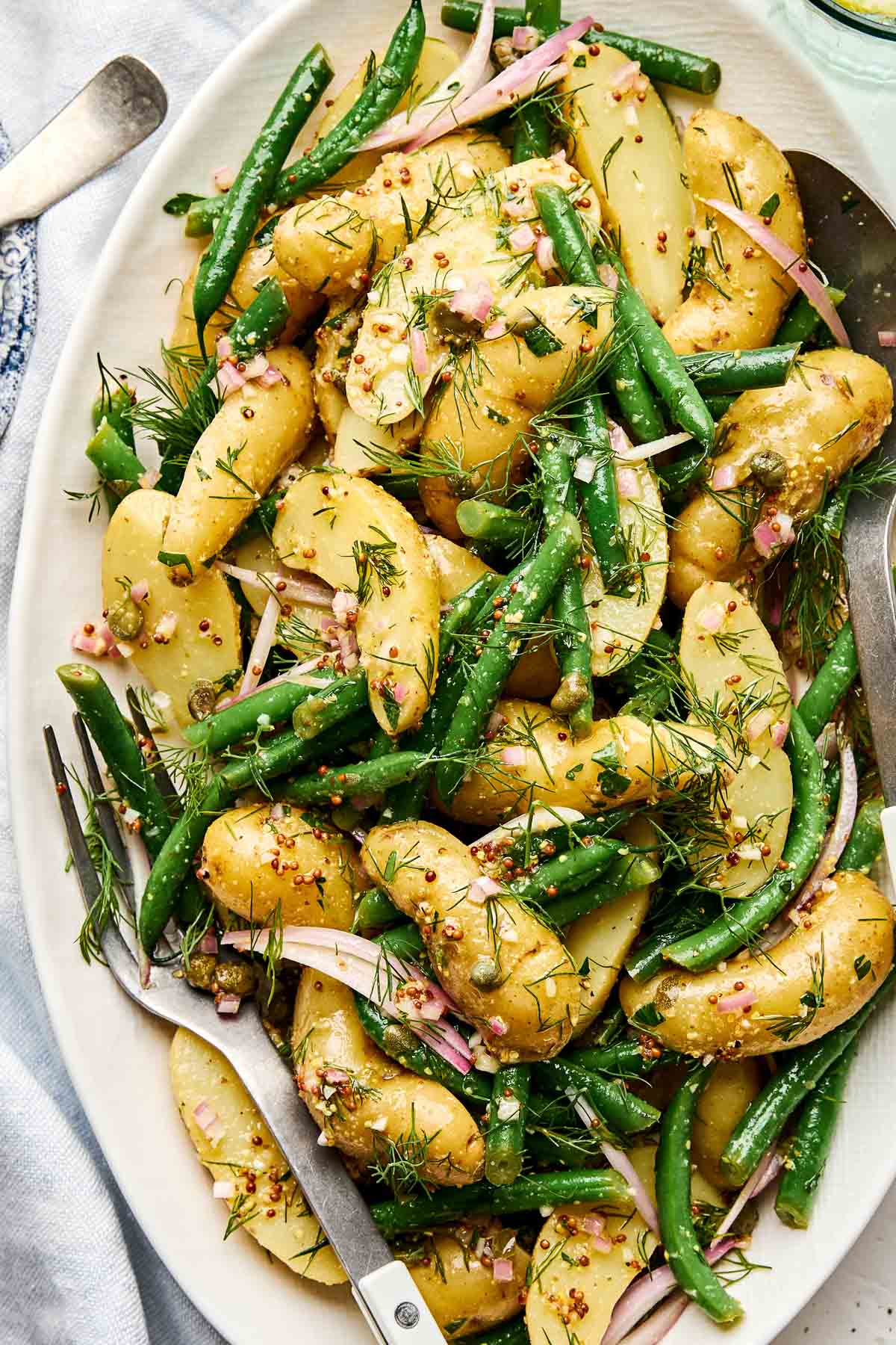A platter of fingerling potatoes, green beans, sliced red onions, capers, and fresh dill, tossed in a mustard vinaigrette. Two serving utensils rest on the side.