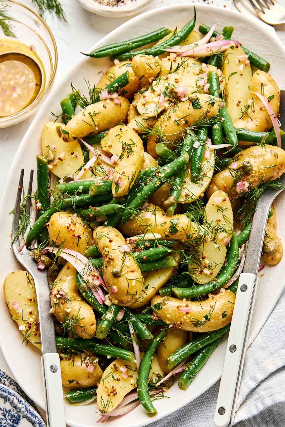 A platter of potato salad with green beans, topped with fresh dill and thinly sliced red onions, served with a mustard vinaigrette and two serving utensils.