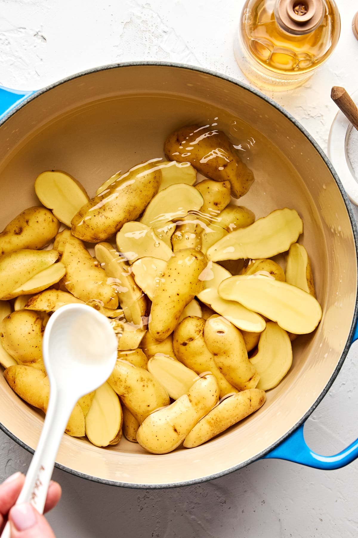 A blue pot filled with whole and sliced fingerling potatoes being drizzled with vinegar, with a hand holding a white spoon above the pot. A bottle of vinegar and a lid are also visible nearby.