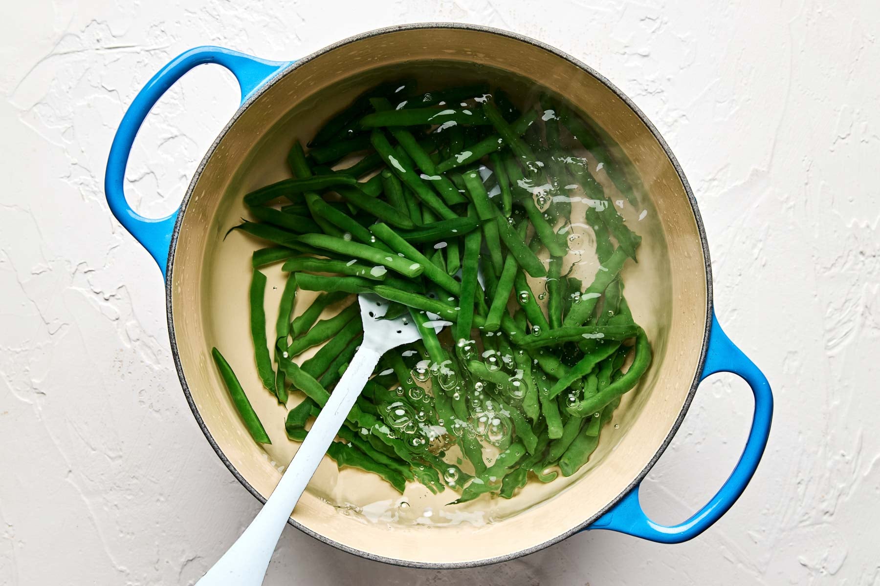 Green beans are being blanched in a blue pot filled with boiling water. A white slotted spoon rests in the pot, stirring the beans. The background is a light textured surface.