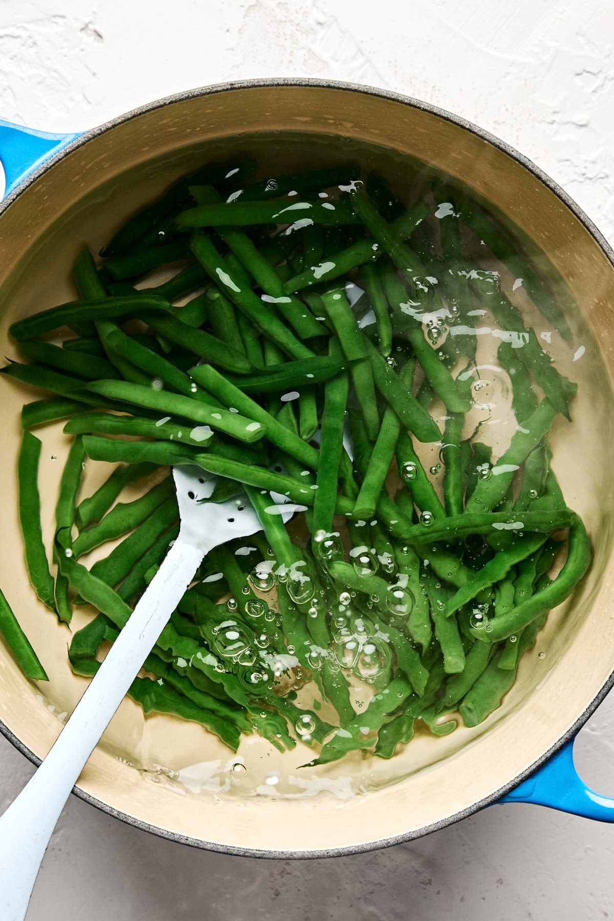 Green beans are being blanched in a pot of boiling water, with a white slotted spoon stirring them. Bubbles are visible around the beans, and the pot has a blue handle.