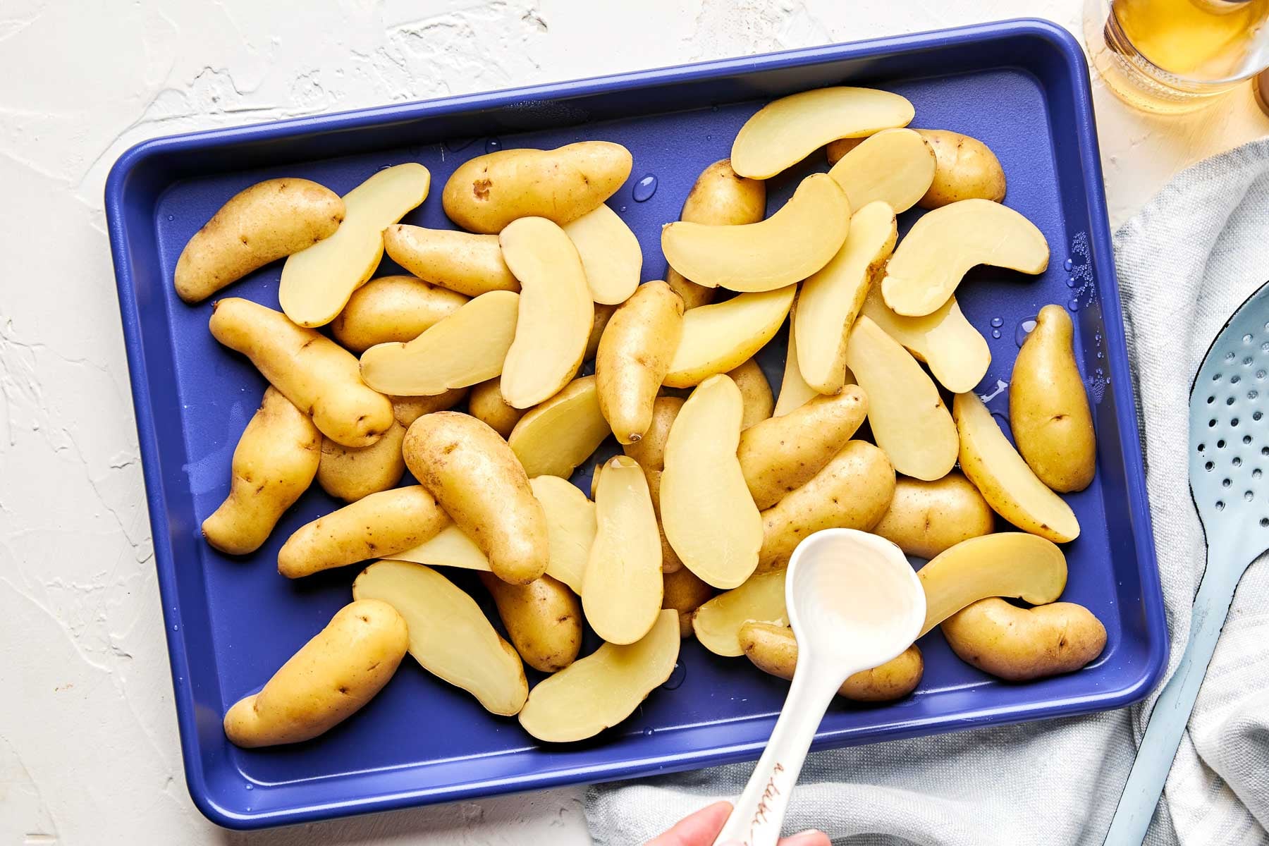 A blue baking tray filled with halved fingerling potatoes, some whole and some sliced, being drizzled with oil from a white measuring spoon. A kitchen towel and slotted spoon are nearby.