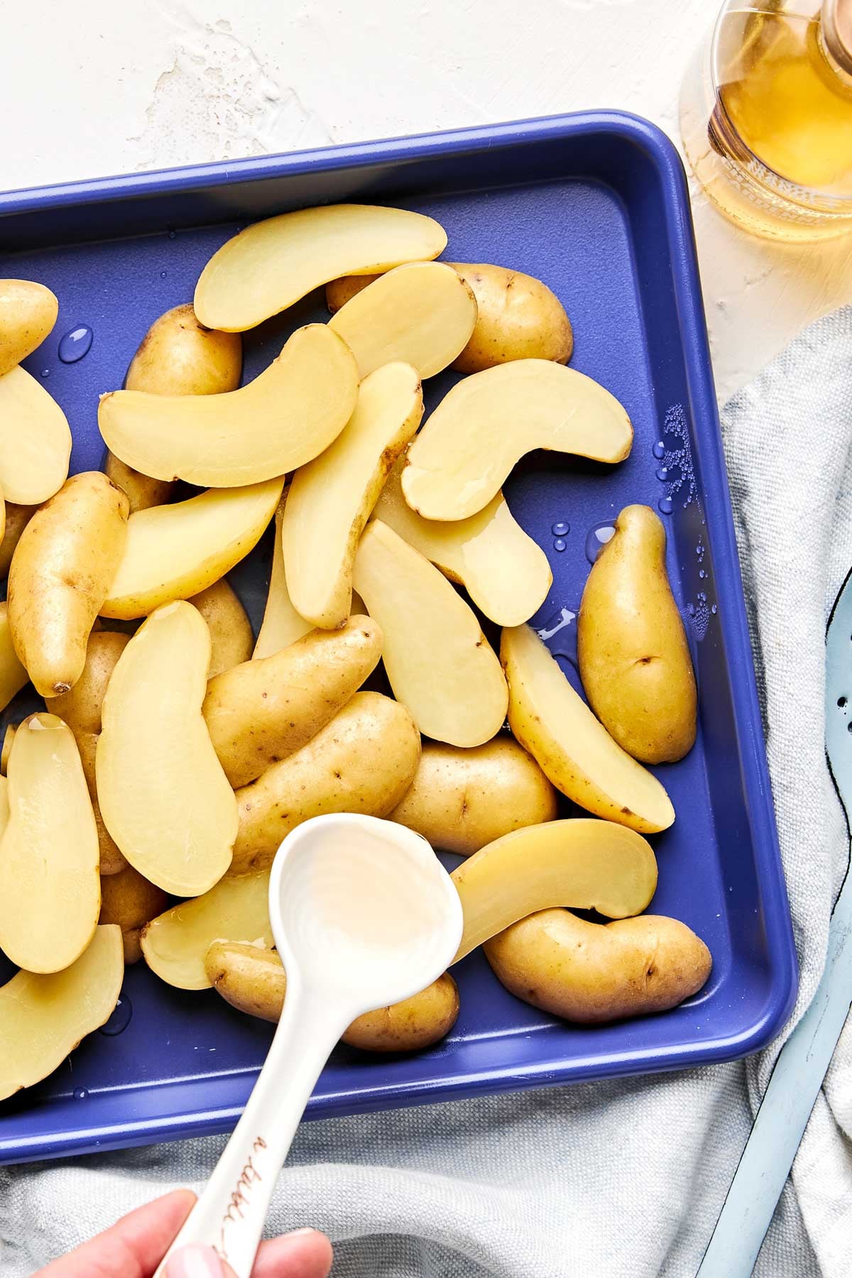 Sliced fingerling potatoes are spread on a blue baking sheet. A white spoon pours vinegar over them. A glass of light-colored liquid sits nearby on a table with a light cloth.