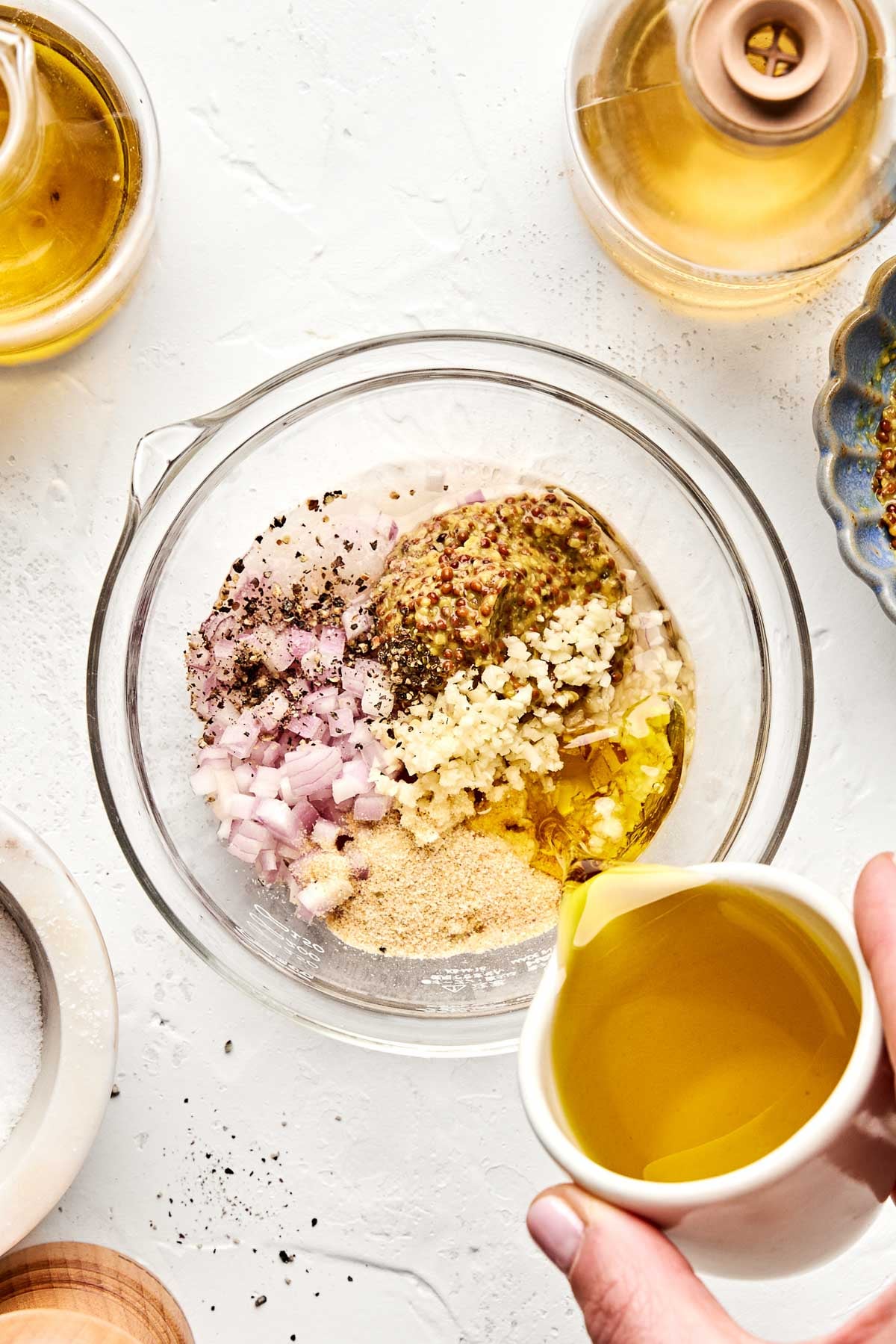 A hand pours olive oil from a small white pitcher into a clear bowl containing chopped shallot, minced garlic, mustard, black pepper, and other seasonings on a white countertop.