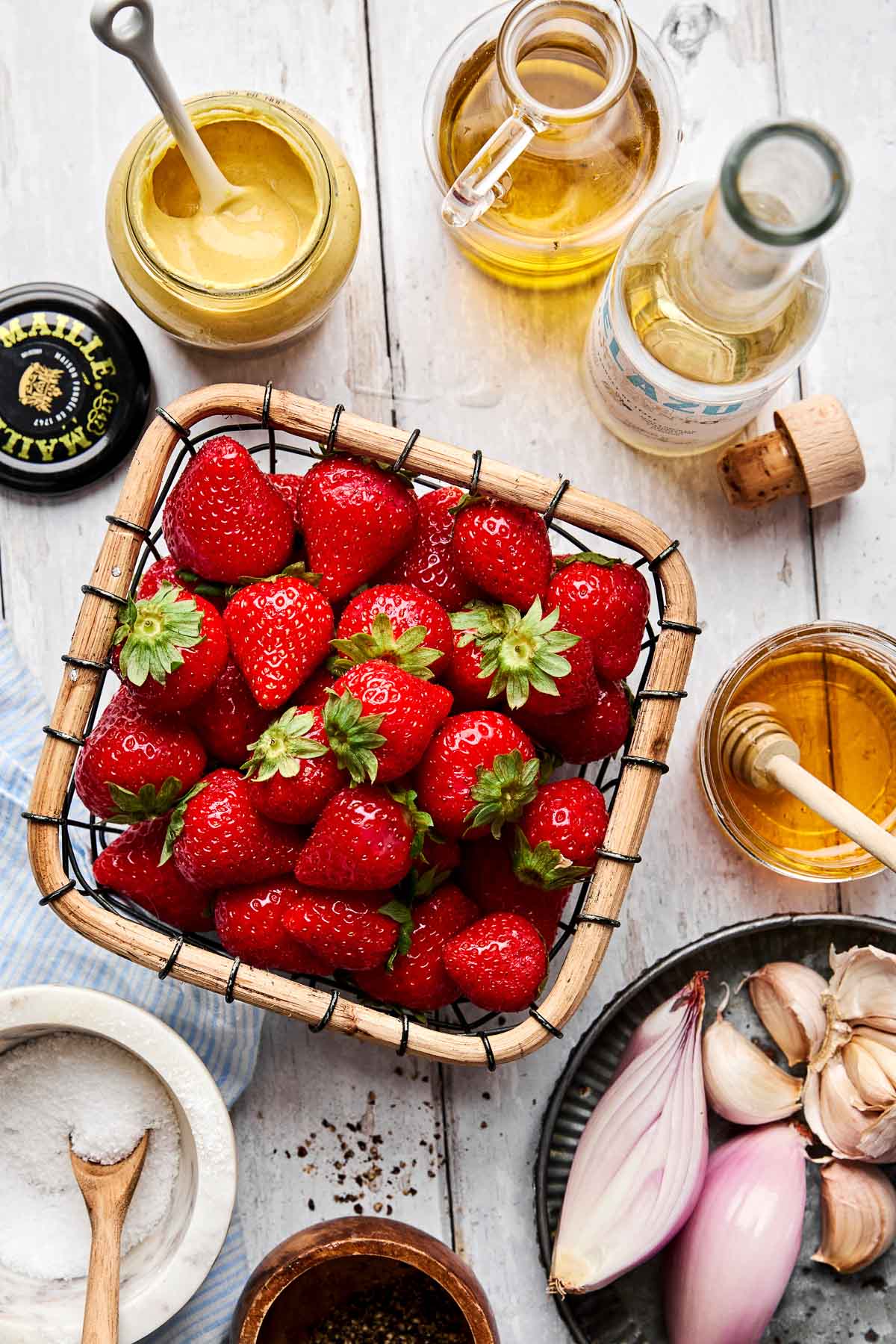 A basket of fresh strawberries surrounded by jars of mustard, vinegar, oil, honey, a bowl of salt, ground pepper, shallots, and garlic cloves on a white wooden surface.
