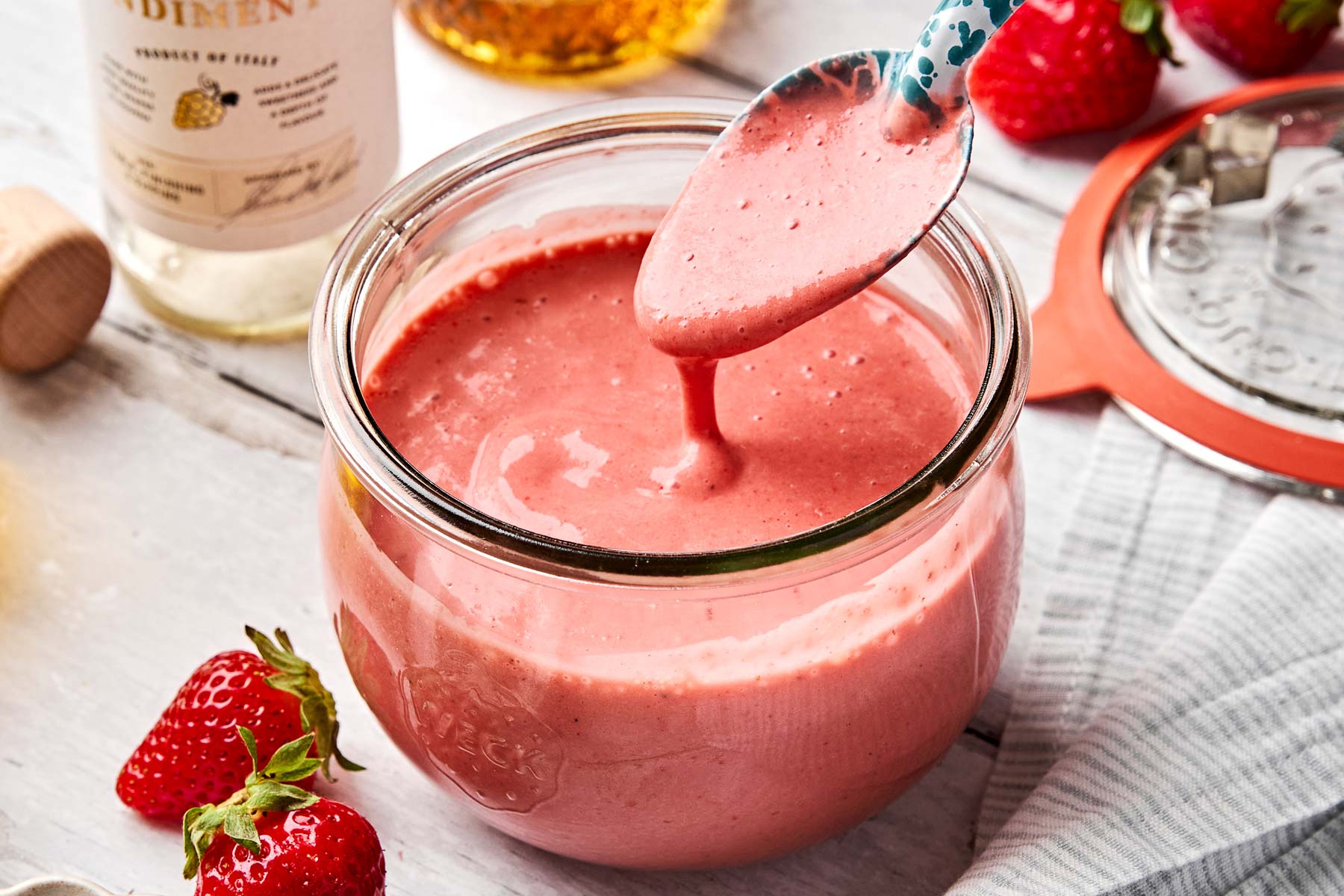 A spoon drips strawberry dressing into a glass jar filled with the pink sauce. Fresh strawberries and a striped towel are nearby on a white wooden surface. Bottles and a jar lid are in the background.
