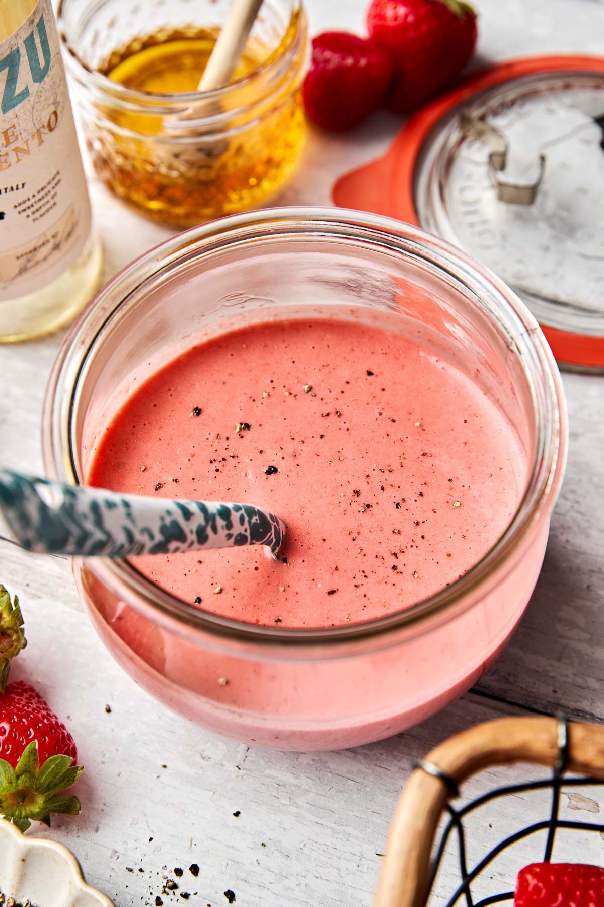 A glass jar filled with pink strawberry vinaigrette, sprinkled with black pepper, with a spoon inside. Surrounding the jar are strawberries, honey, a bottle, and a jar lid on a white surface.