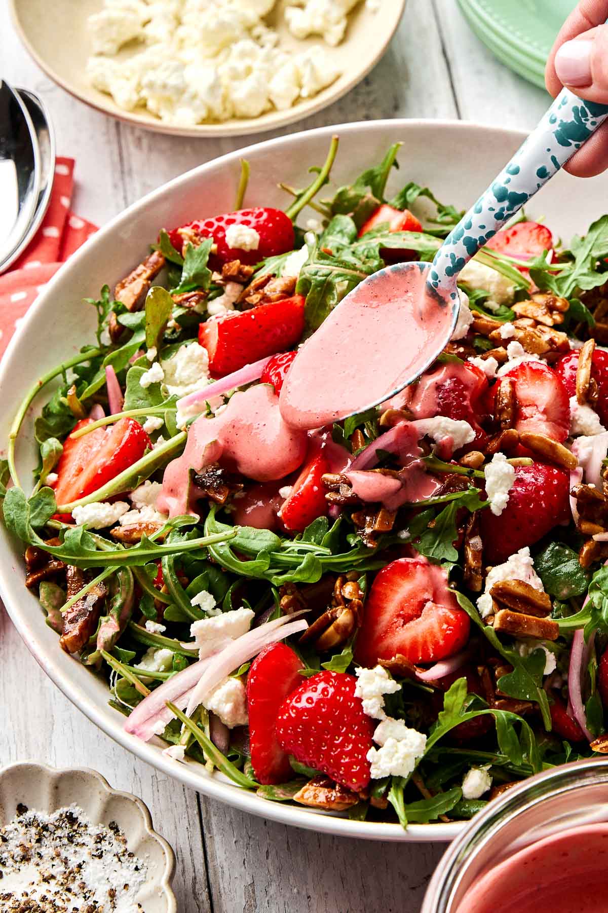 A bowl of fresh arugula salad topped with strawberries, pecans, shallot, and crumbled cheese, being drizzled with pink strawberry dressing from a spoon.