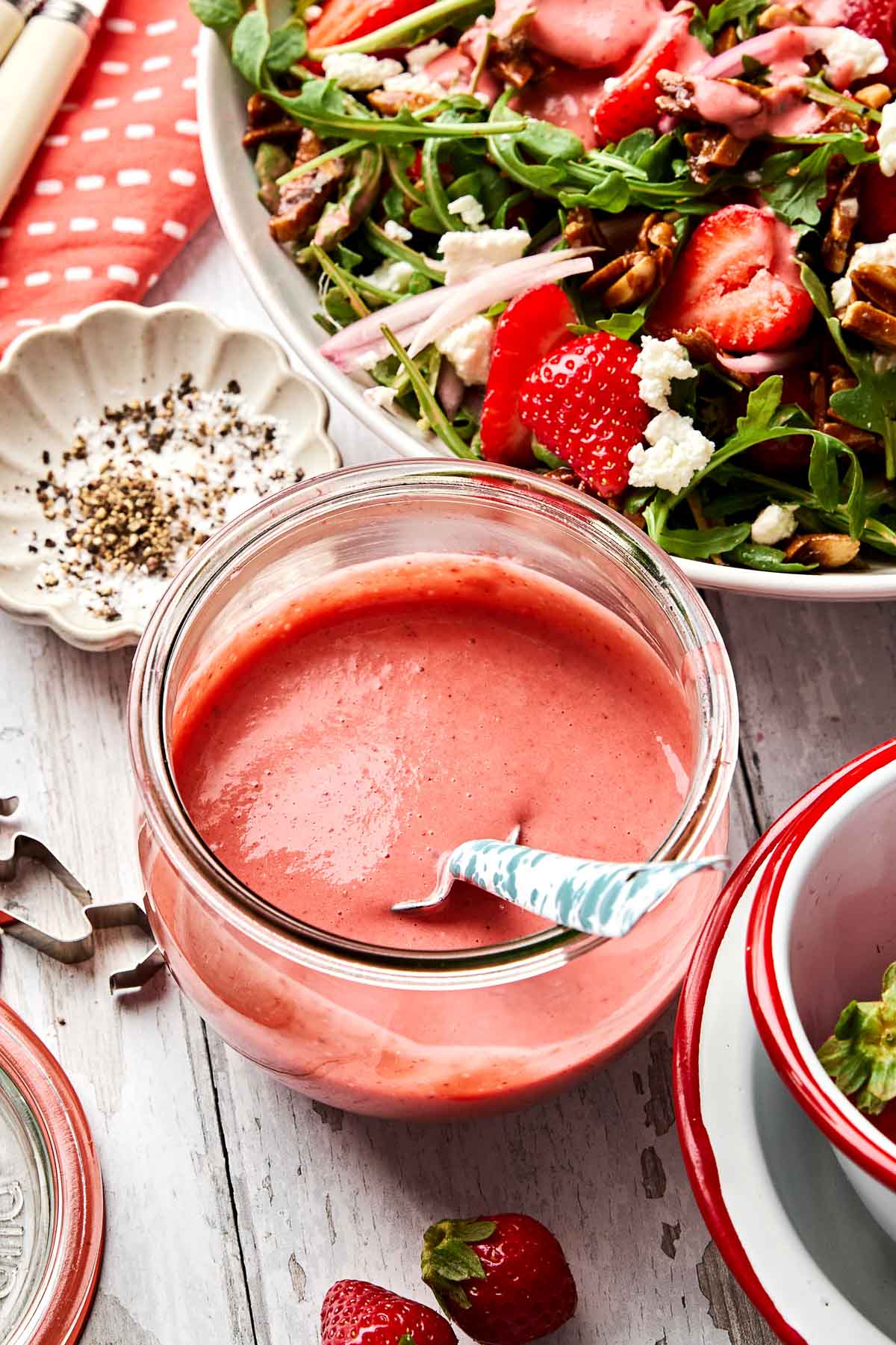 A jar of pink strawberry salad dressing with a spoon sits on a wooden table next to a fresh salad with arugula, strawberries, nuts, and cheese. A small dish with cracked black pepper is nearby.
