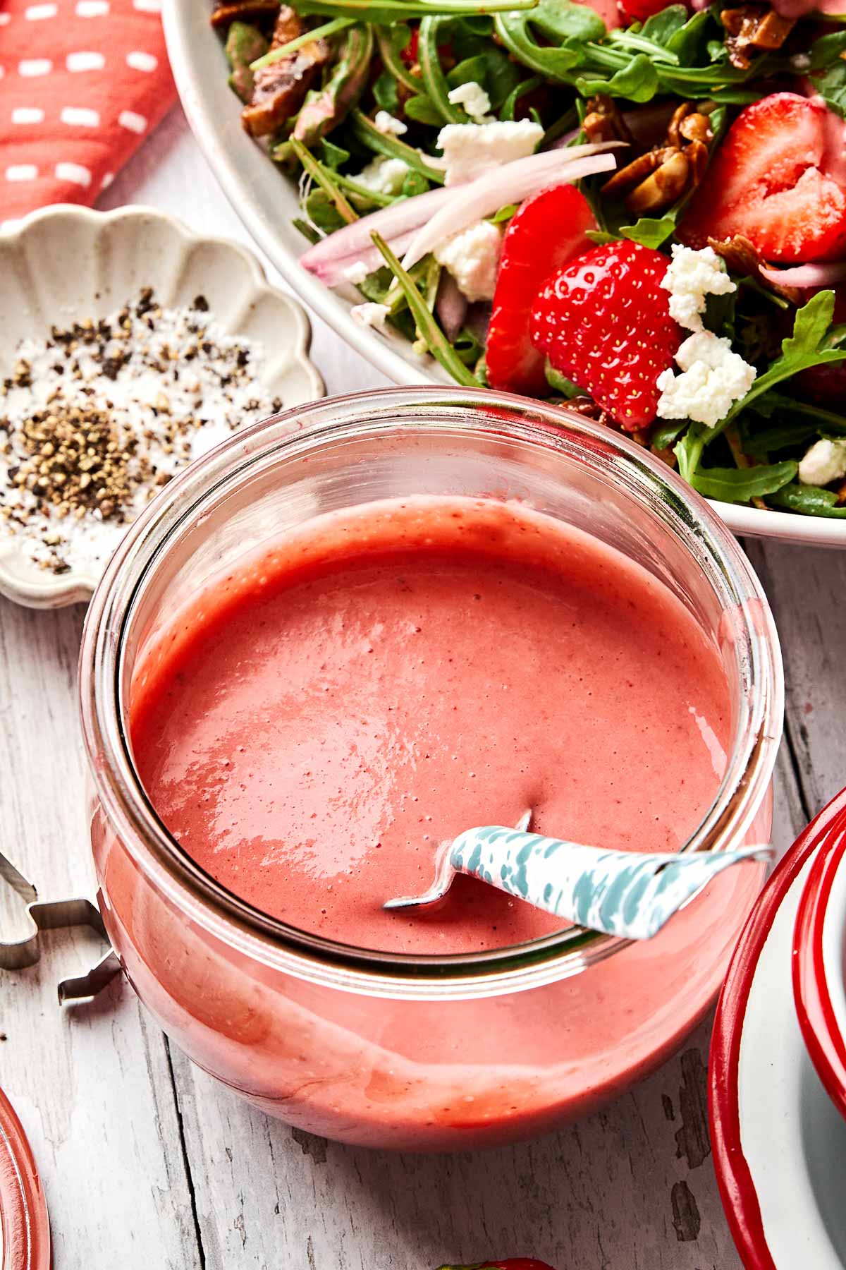 A jar of creamy strawberry balsamic vinaigrette with a spoon in it sits on a white wooden table next to a fresh salad with greens, strawberries, goat cheese, and pecans. A small dish of ground pepper is also nearby.