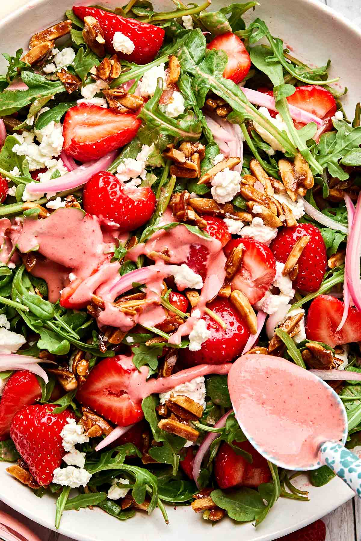 A close-up of a fresh salad with arugula, sliced strawberries, crumbled feta, pickled onions, candied pecans, and pink strawberry vinaigrette being drizzled with a spoon.