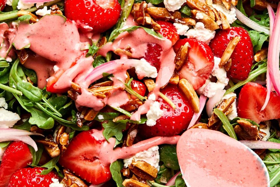 A close-up of a fresh salad with arugula, sliced strawberries, crumbled cheese, pickled onions, glazed nuts, and a pink strawberry dressing drizzled on top. A spoon with more dressing is visible in the corner.