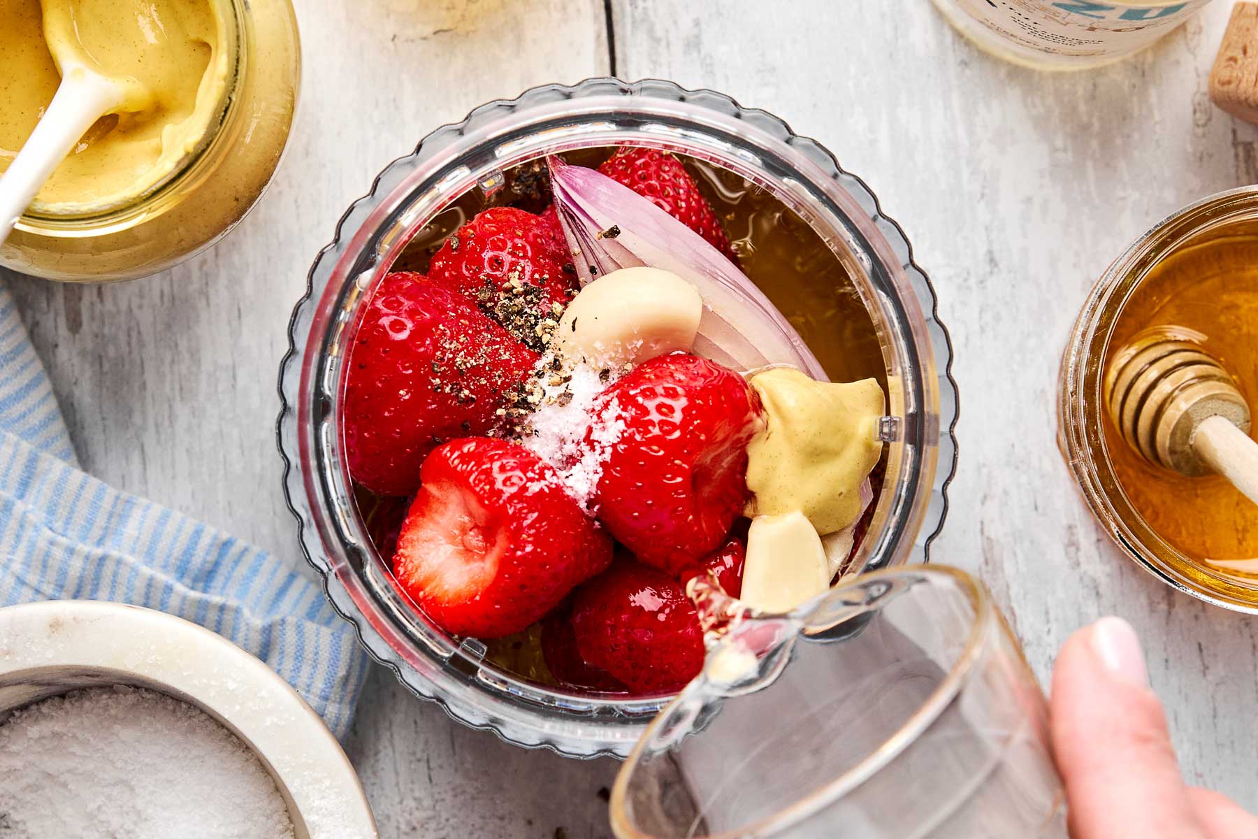 A glass blender cup filled with strawberries, garlic, shallot, mustard, salt, pepper, and honey, with a hand pouring liquid into it. Other jars of mustard and honey are nearby on a white wooden surface.