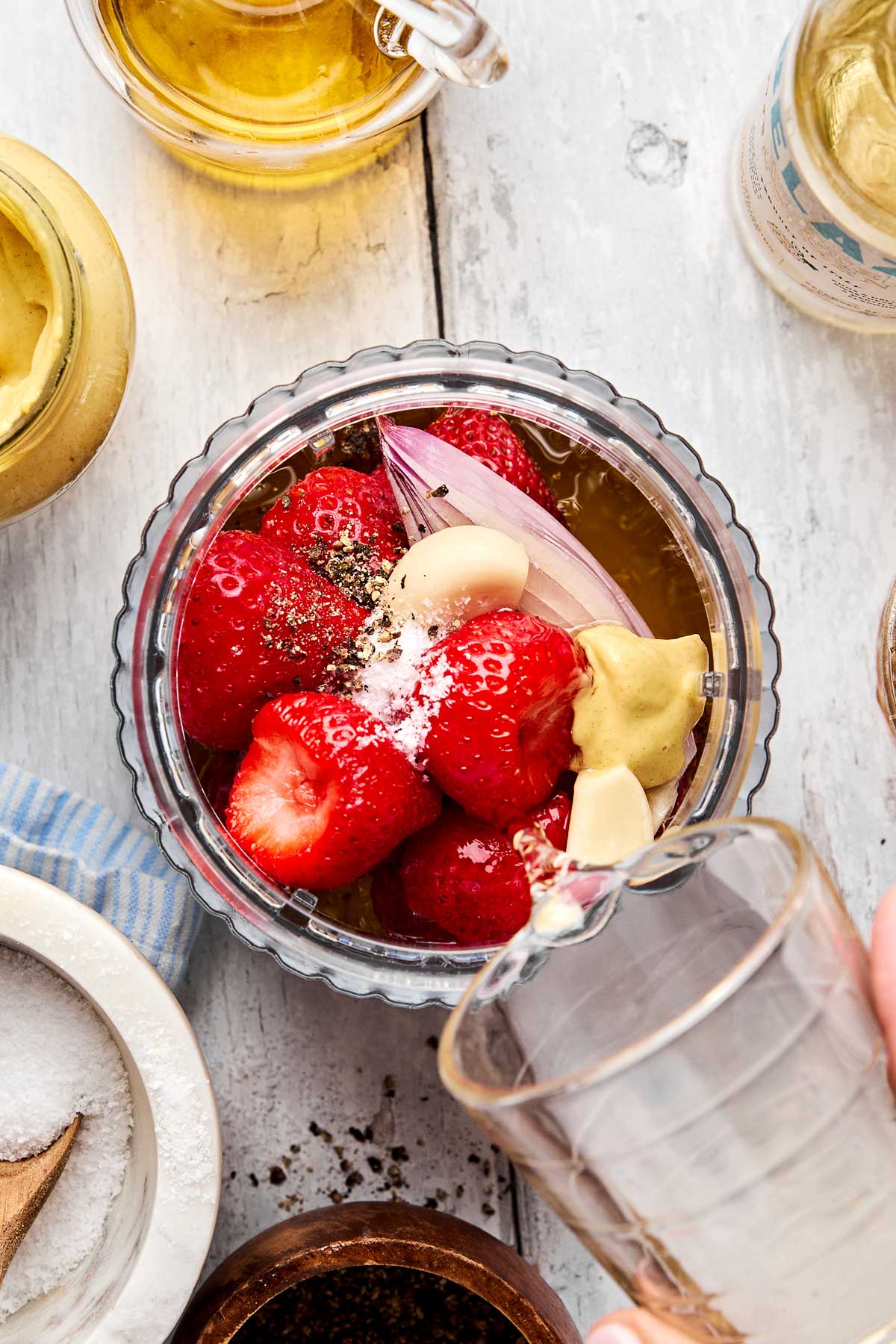 A top-down view of a blender cup filled with fresh strawberries, whole garlic cloves, mustard, a shallot, salt, and pepper, with a hand pouring liquid from a glass into the cup. Other ingredients are arranged around it on a white surface.