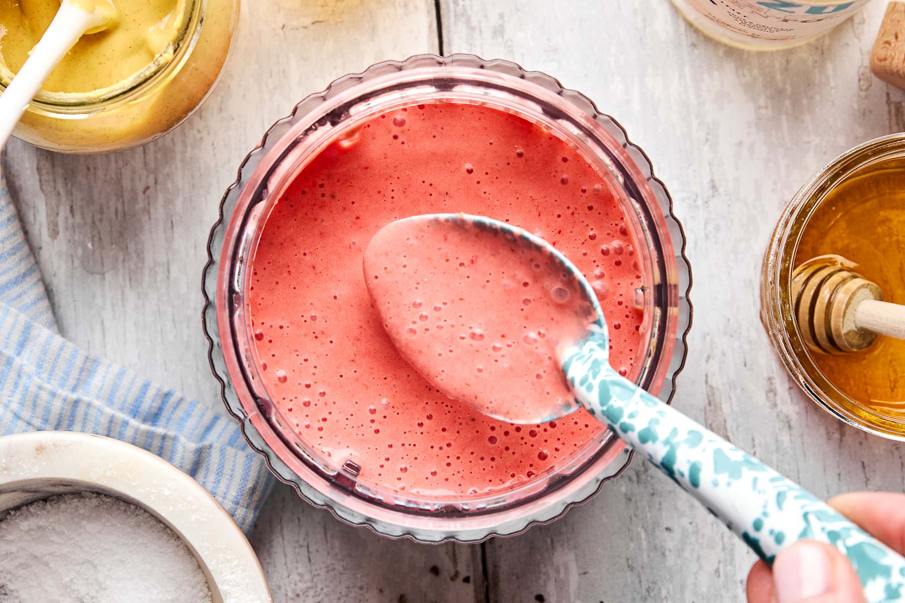 A hand holds a patterned spoon above a glass bowl filled with frothy pink smoothie, surrounded by jars of honey, mustard, and a blue-striped cloth on a white wooden surface.