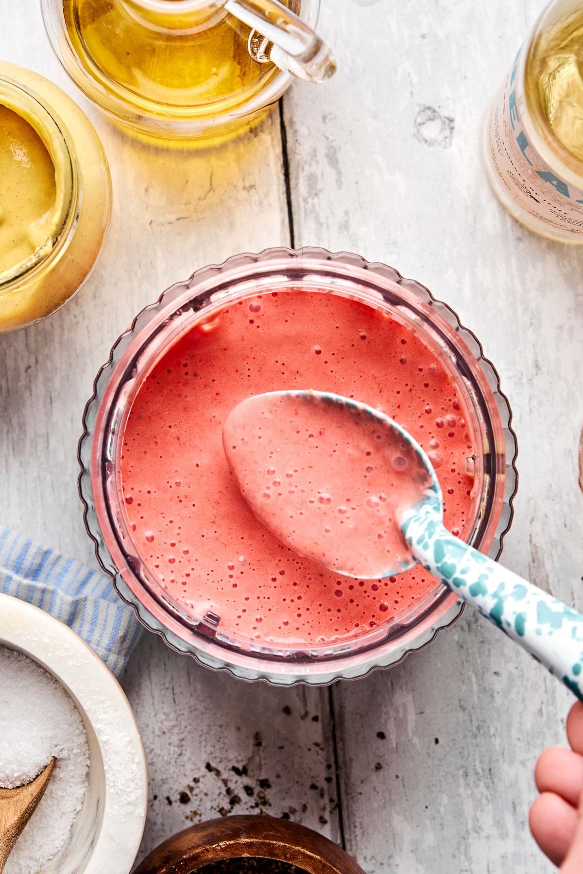 A bowl of pink, frothy salad dressing being stirred with a patterned spoon, surrounded by jars of oil and mustard, a small bowl of salt, and a striped napkin on a white wooden surface.