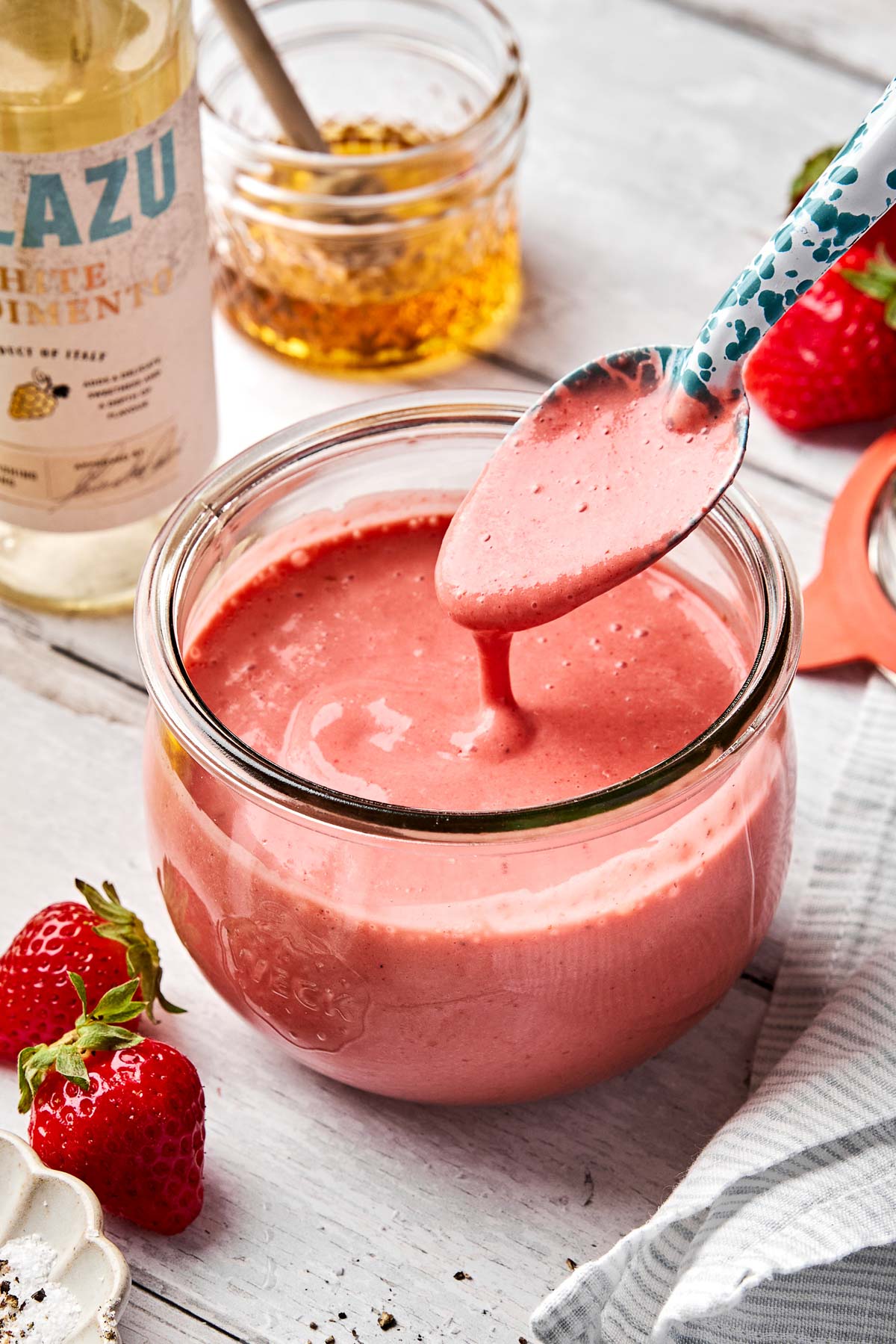 A spoon holds up creamy pink strawberry vinaigrette above a glass jar filled with the dressing. Fresh strawberries, a bottle of white condiment, and a jar of honey are nearby on a light wooden surface.