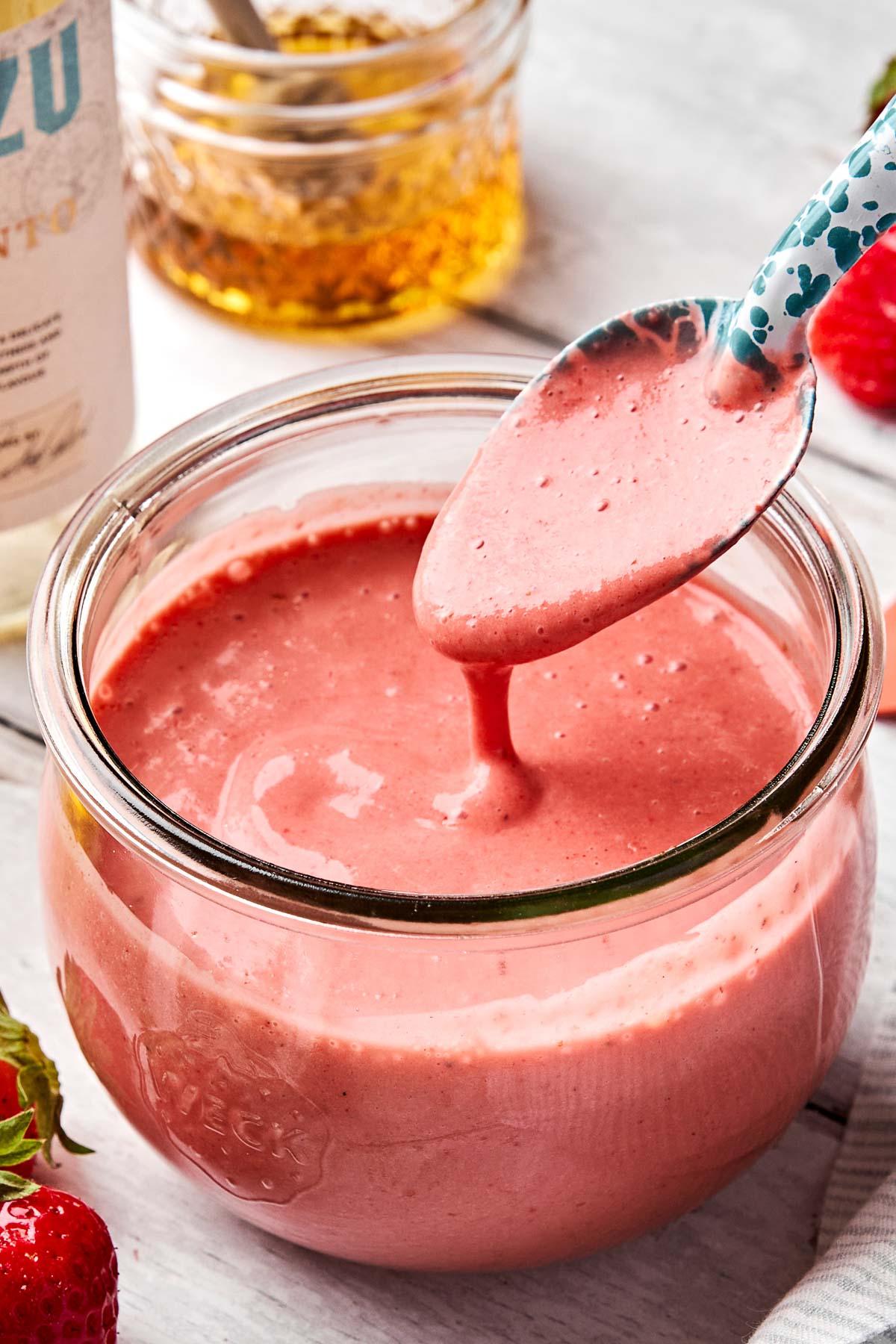 A spoon hovers over a glass jar filled with thick, creamy pink strawberry salad dressing. Fresh strawberries and a jar of honey are visible in the background.