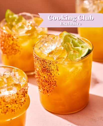 Four glasses of a bright orange drink with ice, lime wedges, and chili powder on the rims are on a pink surface. A text banner reads Cooking Club Exclusive in the upper right corner.