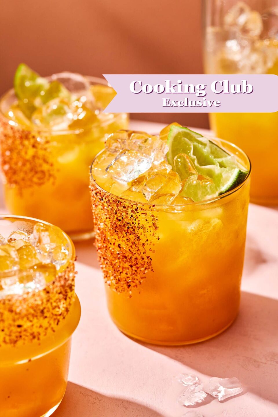Four glasses of a bright orange drink with ice, lime wedges, and chili powder on the rims are on a pink surface. A text banner reads Cooking Club Exclusive in the upper right corner.