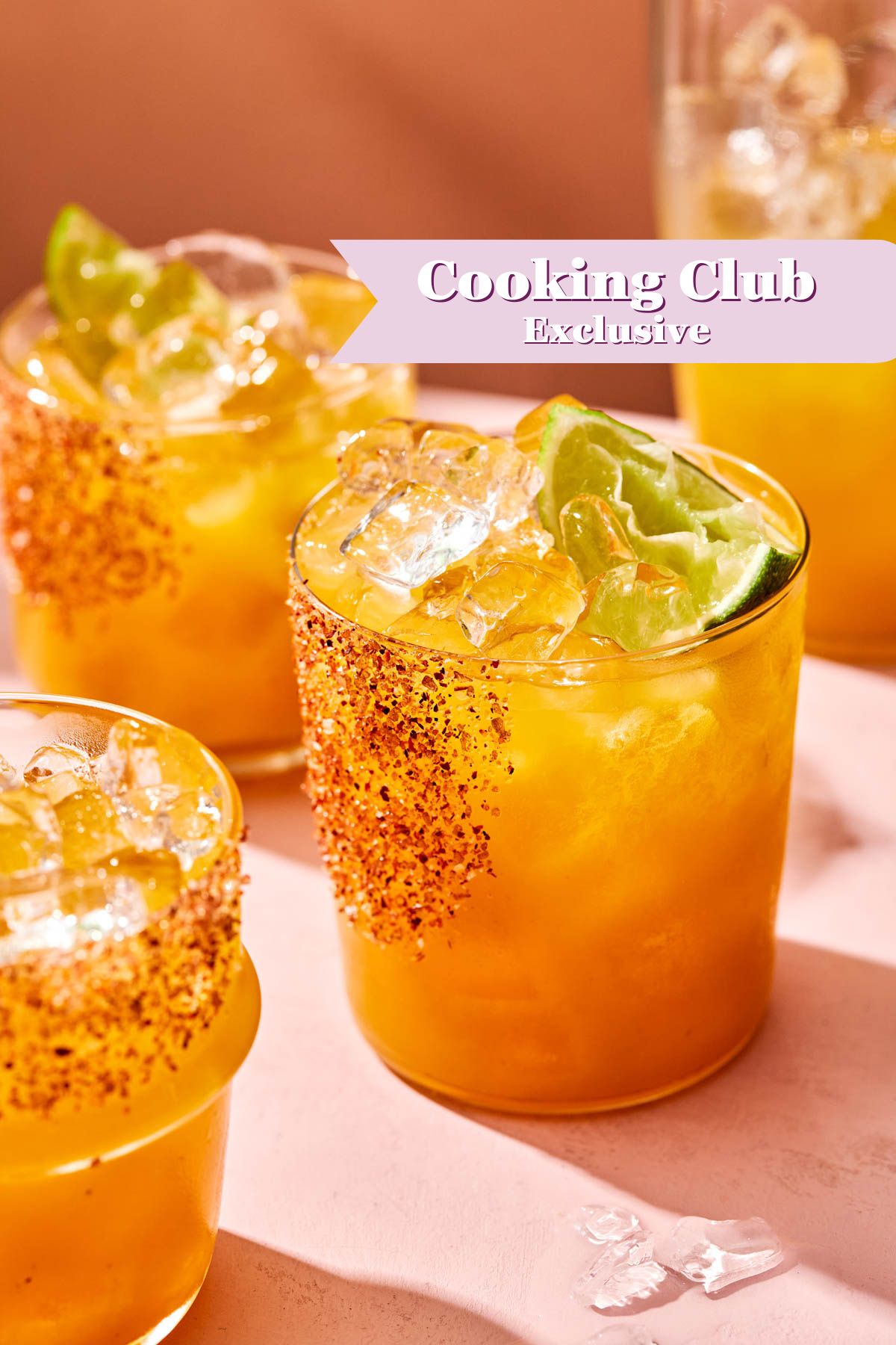 Four glasses of a bright orange drink with ice, lime wedges, and chili powder on the rims are on a pink surface. A text banner reads Cooking Club Exclusive in the upper right corner.