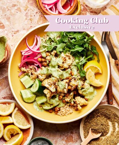 A roasted cauliflower bowl with cucumber, arugula, pickled onions, and green sauce sits on a marble surface, surrounded by lemon wedges, herbs, salt, and a drink. A banner reads Cooking Club Exclusive.