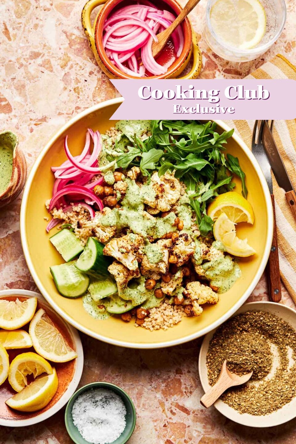A roasted cauliflower bowl with cucumber, arugula, pickled onions, and green sauce sits on a marble surface, surrounded by lemon wedges, herbs, salt, and a drink. A banner reads Cooking Club Exclusive.