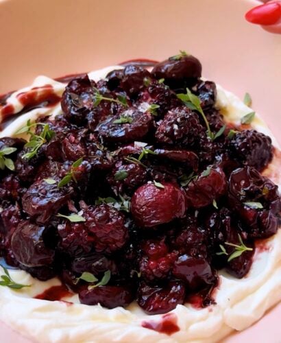 A plate of whipped creamy yogurt topped with a glossy, dark cherry and berry compote, garnished with small fresh thyme leaves.