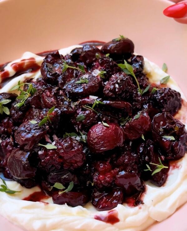 A plate of whipped creamy yogurt topped with a glossy, dark cherry and berry compote, garnished with small fresh thyme leaves.