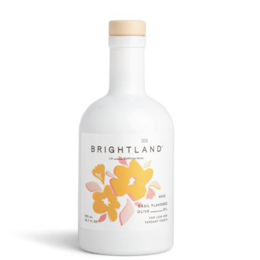 A white bottle of Brightland basil flavored olive oil features a wooden cap and a label with yellow and pink floral illustrations. The text on the bottle indicates it is made in California.