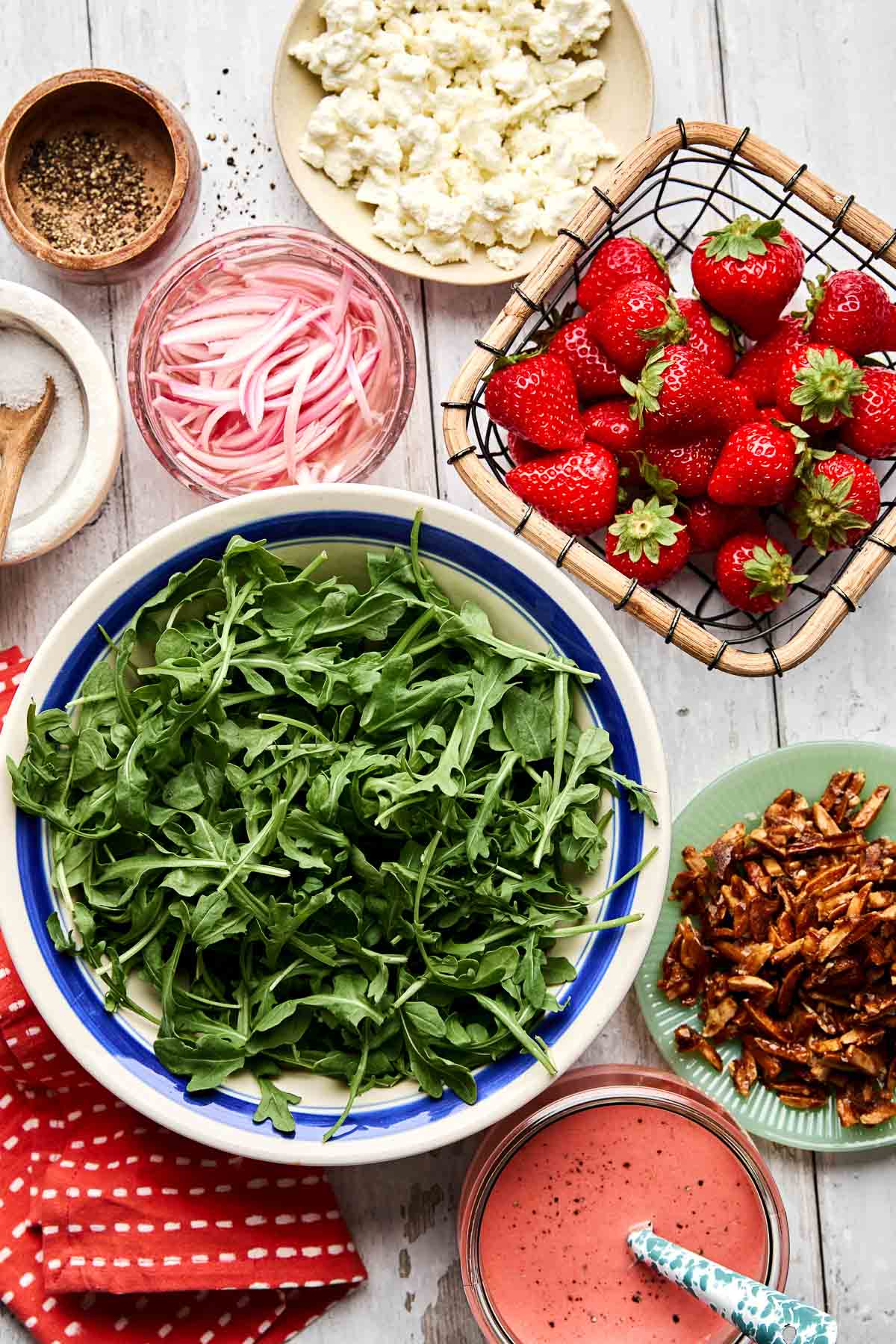 A bowl of fresh arugula surrounded by strawberries in a basket, crumbled cheese, pickled onions, candied nuts, a jar of dressing, pepper, and a red napkin on a light wooden table.