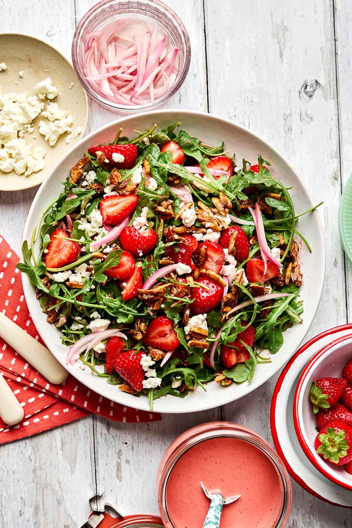 A fresh salad with arugula, strawberries, almonds, goat cheese, and pickled onions in a white bowl. Surrounding the bowl are a small plate of feta, a bowl of pickled onions, utensils, and a jar of pink salad dressing.