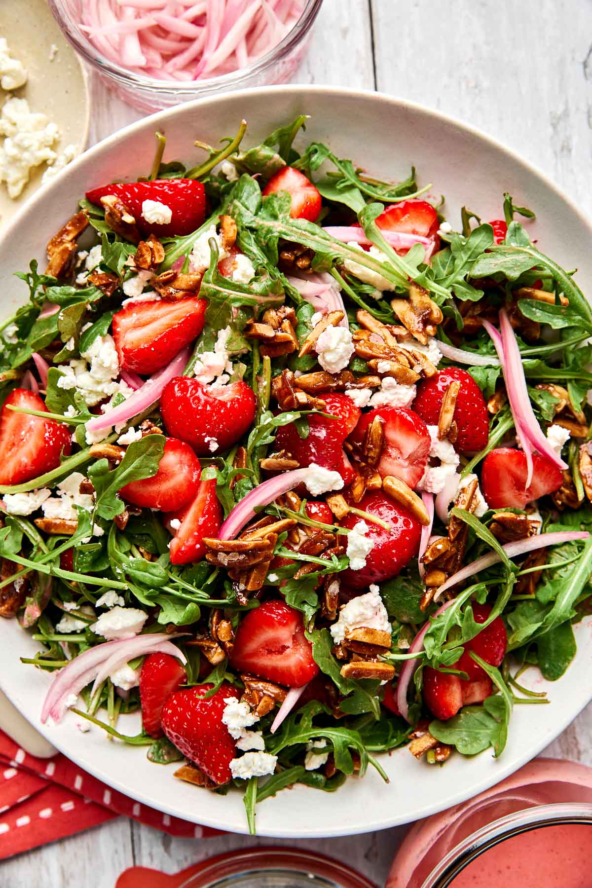 A fresh salad with arugula, sliced strawberries, crumbled goat cheese, candied almonds, and thinly sliced red onions on a white plate. Glasses with pickled onions and strawberry dressing are nearby.