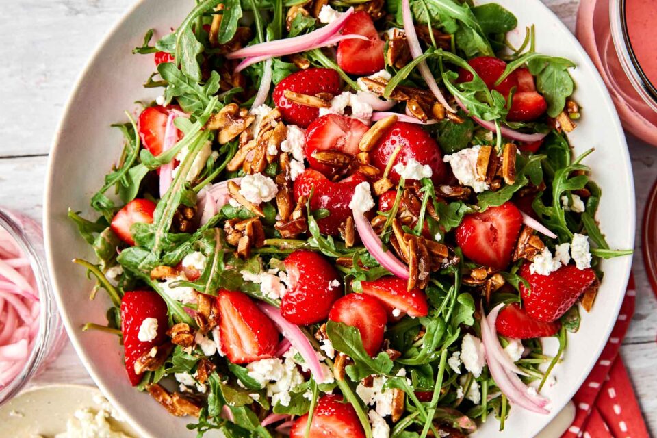 A bowl of fresh salad with arugula, sliced strawberries, crumbled cheese, candied almonds, and pickled red onions on a light background.