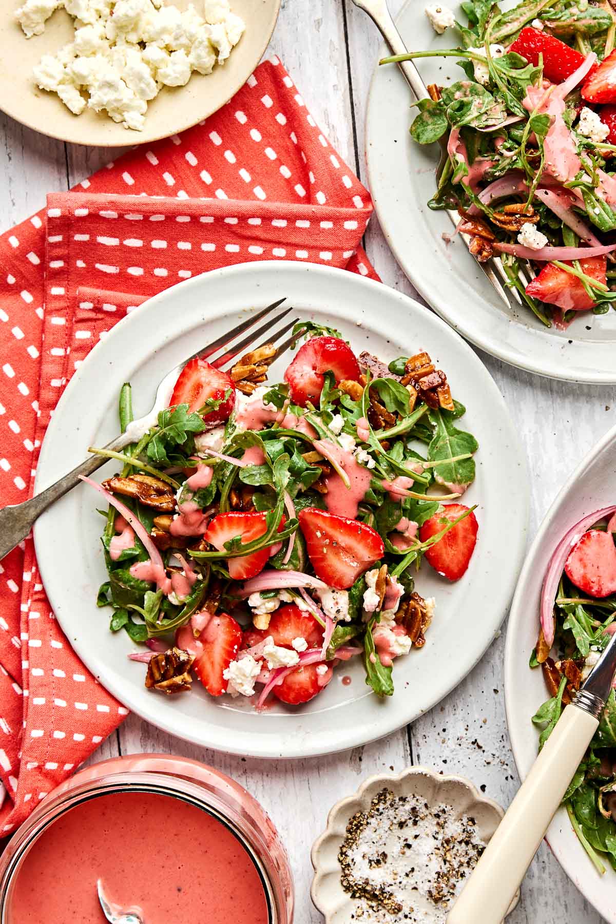 A fresh salad with strawberries, crumbled goat cheese, almonds, and greens is served on a white plate with a fork. A jar of pink dressing, a bowl of feta, and a red dotted napkin are nearby on a white wooden table.